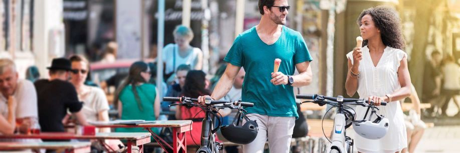 Man en vrouw lopen met fietsen in hand langs het terras terwijl ze in de andere hand beiden een ijsje hebben en met elkaar praten