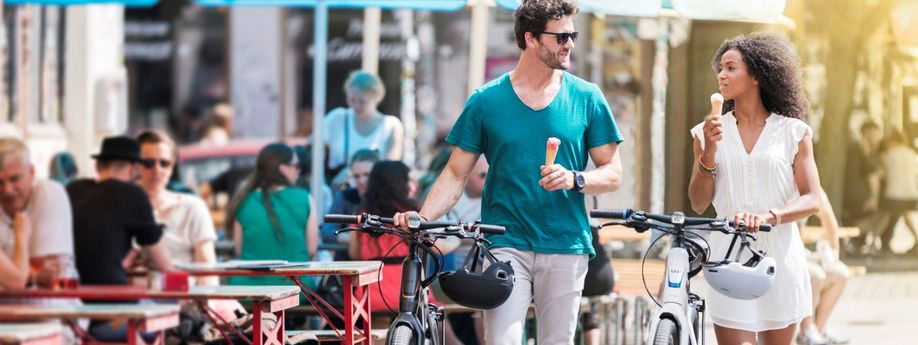 Man en vrouw lopen met fietsen in hand langs het terras terwijl ze in de andere hand beiden een ijsje hebben en met elkaar praten