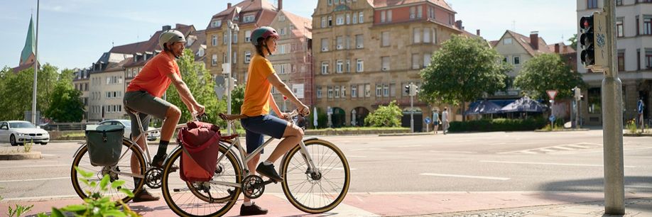 Twee fietsers met beide een gekleurde fietstas voor het rode fietsstoplicht in een stad met unieke huizen op de achtergrond