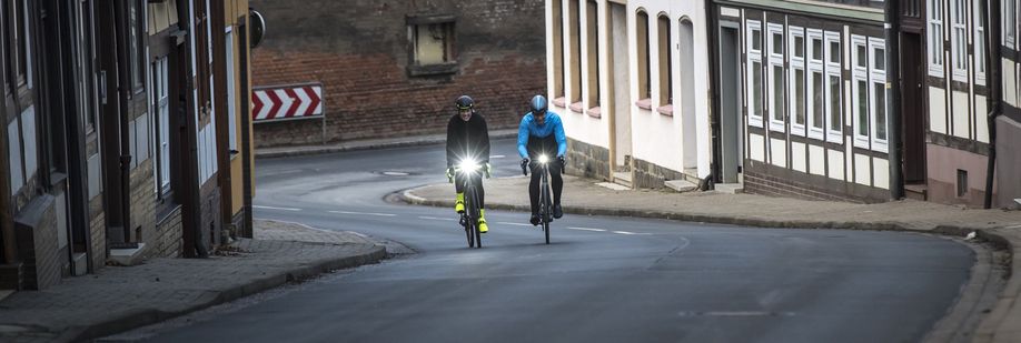 Twee racefietsers met felle voorlichten die een slingerend straatje omhoog fietsen tijdn een koude winterdag