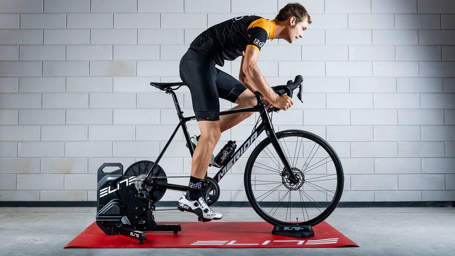 Elite Suito-T fietstrainer op een rode Elite trainermat met Merida fiets erop gemonteerd en persoon die uit het zadel komt met 12GO Biking fietskleding