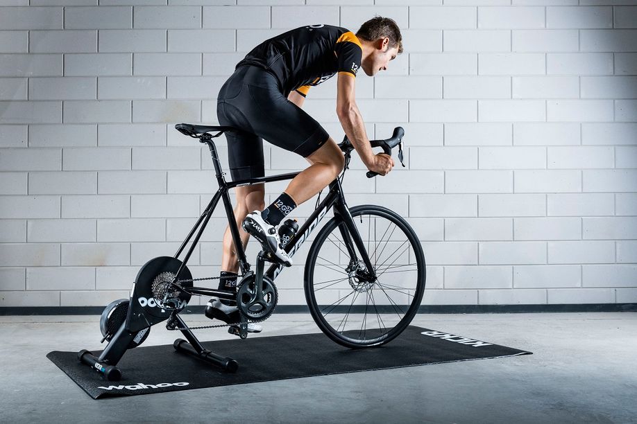 Fietser uit het zadel met Merida fiets op Wahoo Kickr Core en zwarte Wahoo trainermat voor een betongetegelde muur