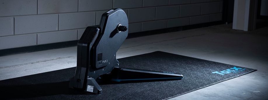 Tacx Flux 2 fietstrainer op een zwarte Tacx trainermat voor een betongetegelde muur