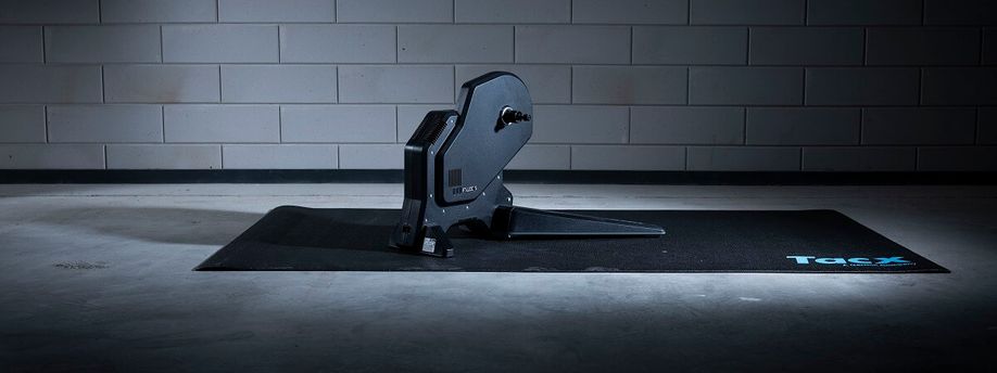 Tacx Flux S fietstrainer op zwarte Tacx trainermat met betongetegelde muur op de achtergrond