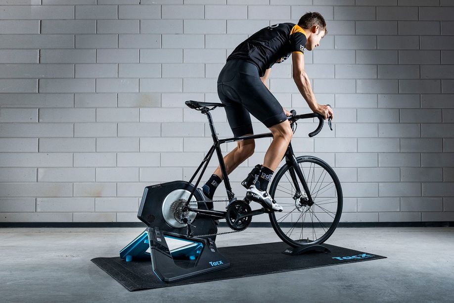 Fietser met 12GO Biking kleding op Merida Racefiets en Tacx Neo 2T fietstrainer op een zwarte Tacx trainermat voor een betongetegelde muur