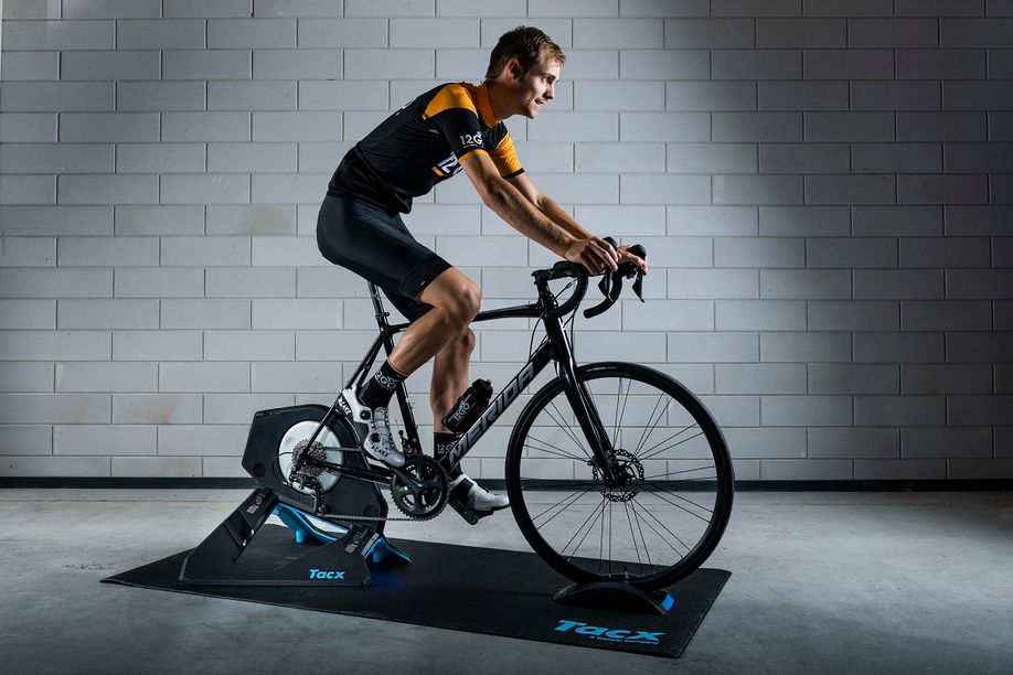 Fietser met 12GO Biking kleding op Merida Racefiets en Tacx Neo 2T fietstrainer op een zwarte Tacx trainermat voor een betongetegelde muur