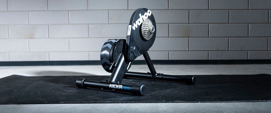 Wahoo Kickr Core fietstrainer op een zwarte Wahoo trainermat met betongetegelde muur als achtergrond