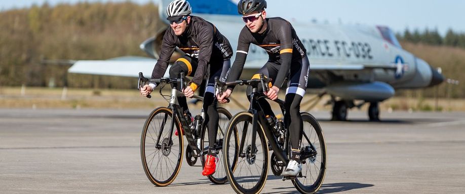 Twee fietsers met 12GO Biking kleding op aero carbon racefietsen met een oude straaljager op de achtergrond