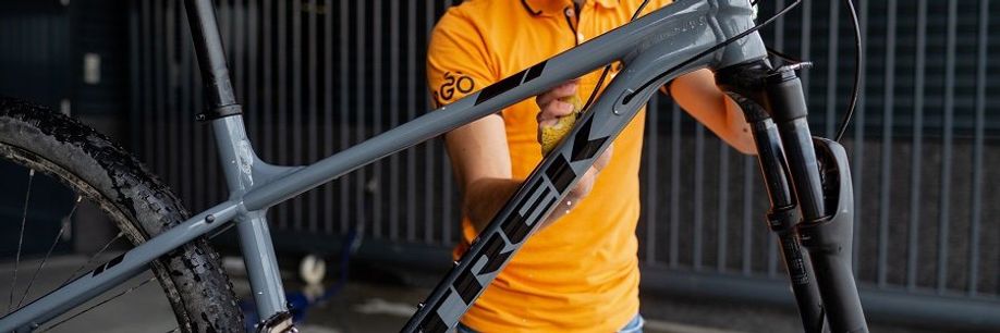Persoon met 12GO Biking shirt maakt het grijze frame van een Trek mountainbike schoon met een gele spons