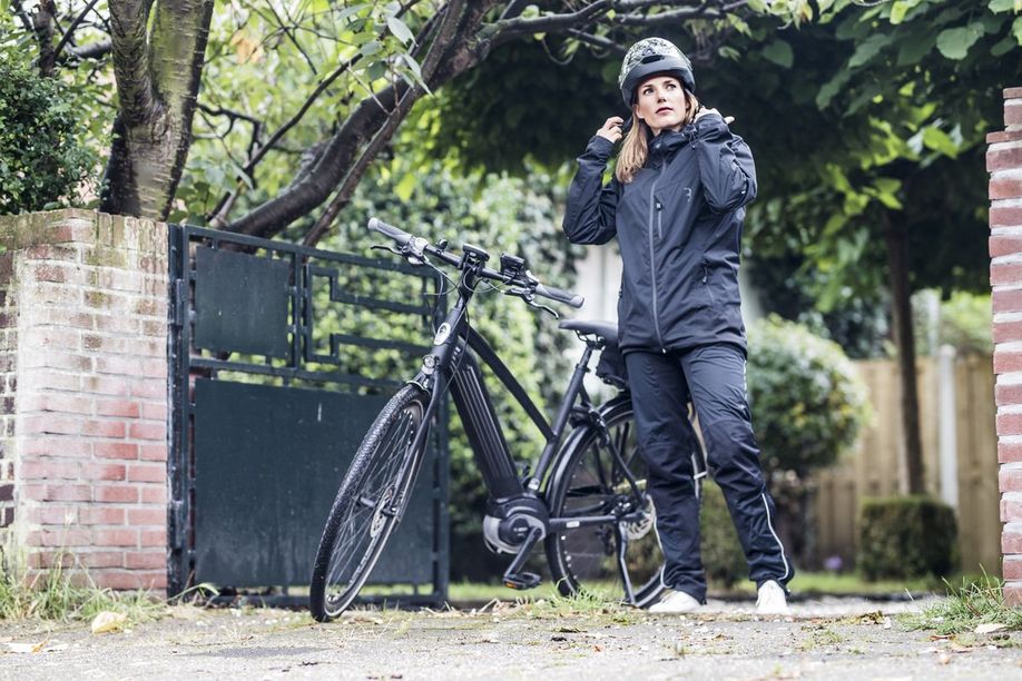 Vrouw met zwart regenpak zet helm op naast haard zwarte elektrische Gazelle e-bike met middenmotor en accu in frame