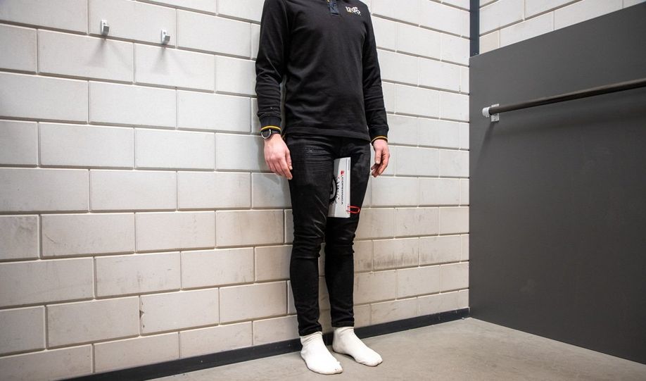 Persoon op sokken recht staand tegen een muur met een boek tussen zijn bovenbenen en tegen zijn kruis om de binnenbeenlengte te meten