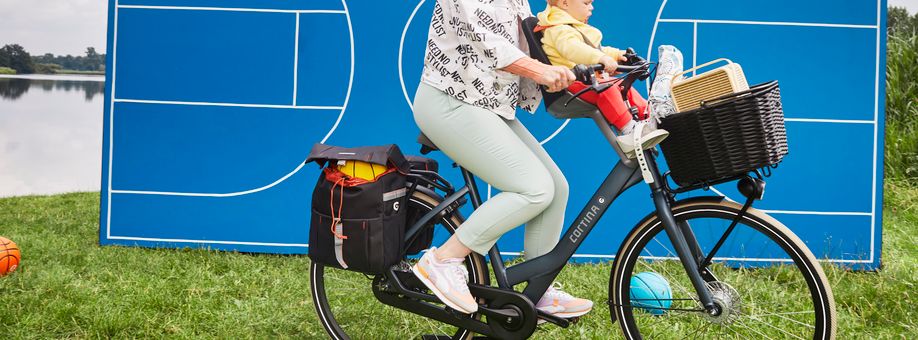 Vrouw met volgeladen fietstassen, fietsmand en kinderzitje met kind op een Cortina fiets in het gras met een blauwe sportveldvlak erachter