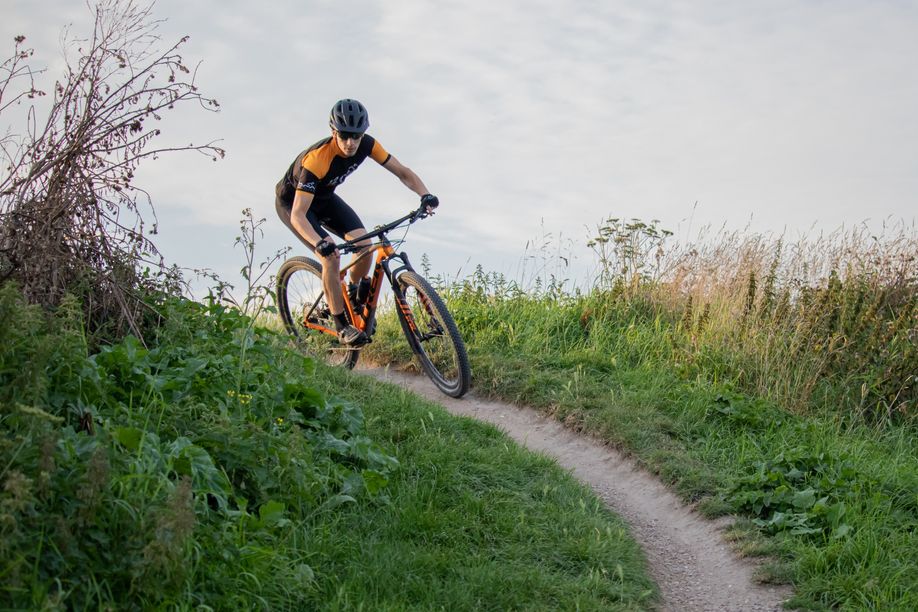 Fietser met oranje Trek MTB en 12GO Fietskleding die een smal paadje afdaalt tussen groen gras en planten