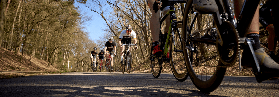 Groep fietsers met 12GO Biking fietskleding die achter elkaar aan sprinten op een asfaltweg tussen de droge bomen door