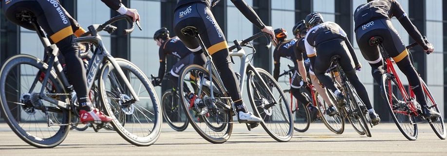 Groep racefietsers met 12GO Biking fietskleding die allemaal een bocht naar links maken over het asfalt voor een gebouw met veel glas