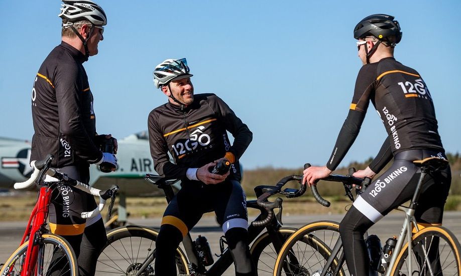 Drie fietsers met 12GO Biking fietskleding die pauzeren met hun racefietsen op luchtbasis Soest met een oud vliegtuig erachter
