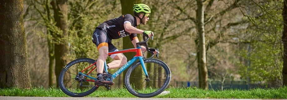 Racefietser met 12GO Biking fietskleding en rood met blauwe Wilier racefiets hangt in de bocht met gras en bomen erachter