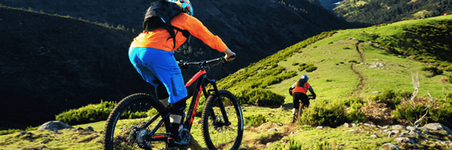 Mountainbiker met Trek MTB en oranje jas met blauwe broek en rugtas volgt persoon met rood shirt op de MTB over een trail door de groene bergen