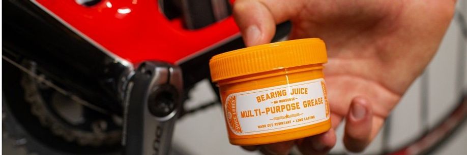 Persoon houdt potje Bearing Juice vast met Dura-Ace crank en rood met zwarte fiets op de achtergrond