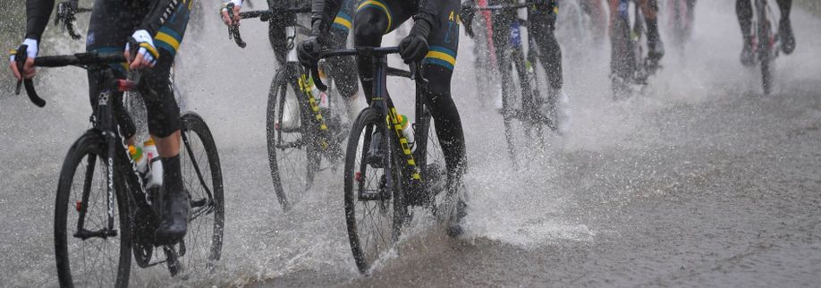 Groep professionele wielrenner die met hun fietsen door een dikke laag water heen fietsen waarbij het water alle kanten op gaat