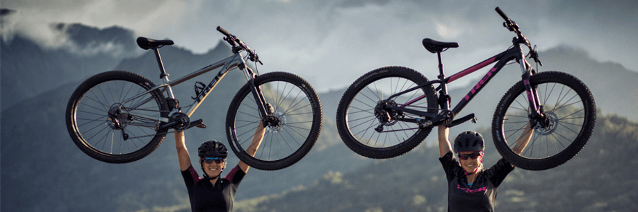 Twee vrouwelijk fietsers houden Trek mountainbikes boven hun hoofd met hoge bergen en wolken op de achtergrond