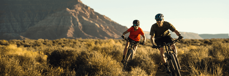 Mannelijke en vrouwelijke mountainbikers rijden op woestijnpad door de struiken met roodachtige bergen op de achtergrond