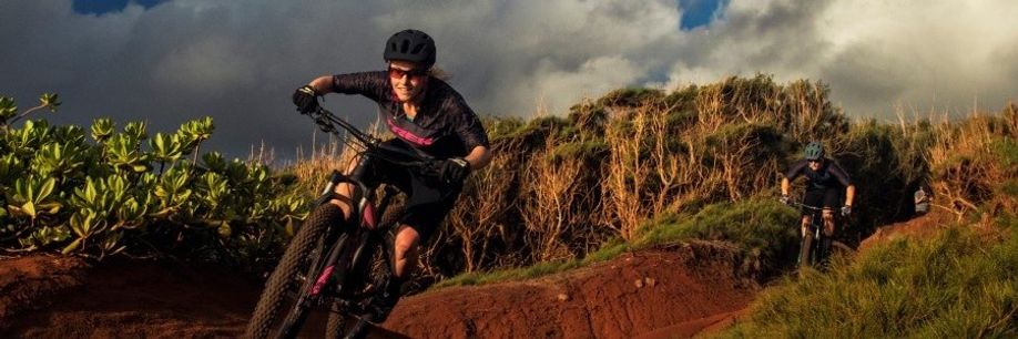 Vrouwelijke mountainbiker met Trek fietsshirt en MTB die een trail afdaalt met een man op de achtergrond die haar niet bij houd