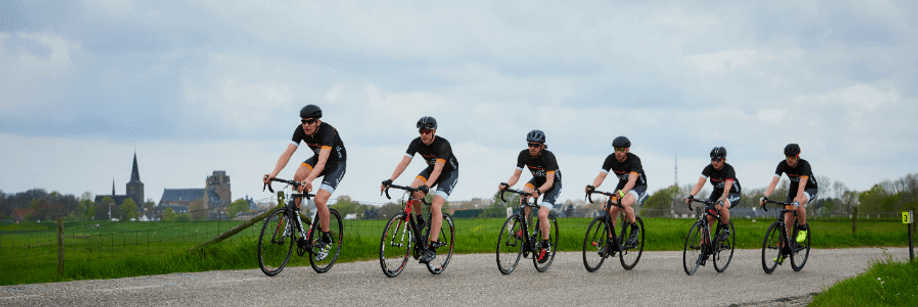 Groep wielrenners met 12GO Biking fietskleding die over een dijkje tussen de weilanden rijden met een dropje in de achtergrond