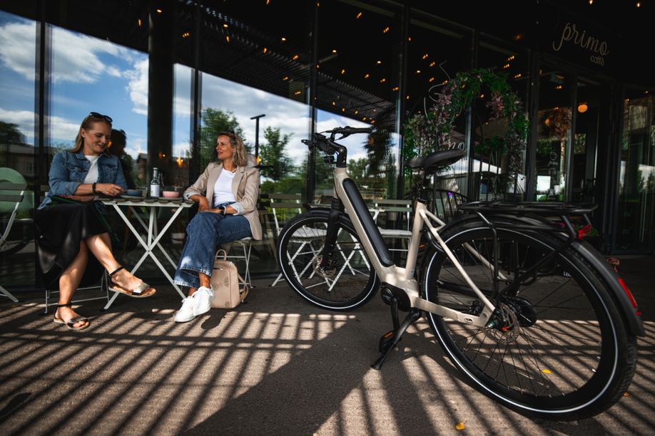 Beige Superior e-bike in de schaduw voor een restaurantje met twee vrouwen die koffie drinken op het terras