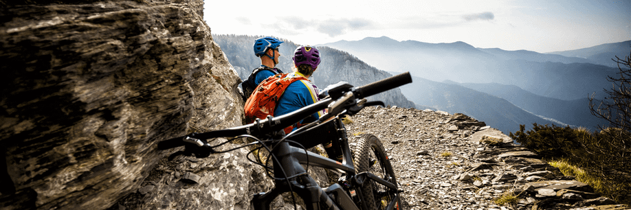 Twee mountainbikers zitten op de zijkant van een bergpad met afgrond en kijken uit over het dal terwijl een mountainbike naast hun tegen de rots aanstaat