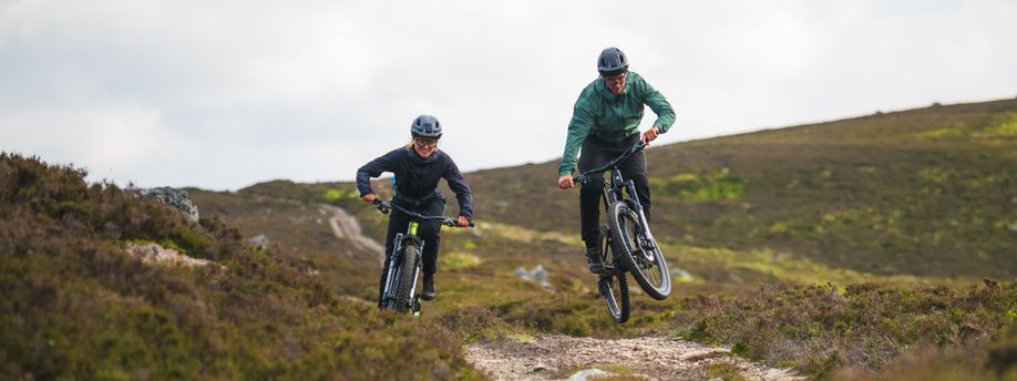 Man en vrouw zijn aan het mountainbike in de heuvels terwijl ze afdalen en de man een sprong maakt op de trail die ze fietsen