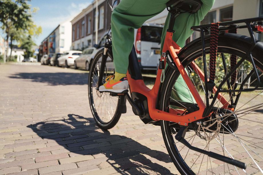 Vrouw met Nike schoenen op een oranje elektrische Sparta fiets op een klinkerstraat met geparkeerde auto's voor de huizen