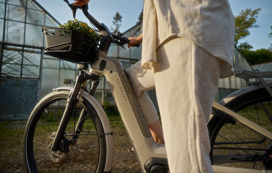 Vrouw met beige e-bike en plantje voorop op het gras voor een aantal kassen