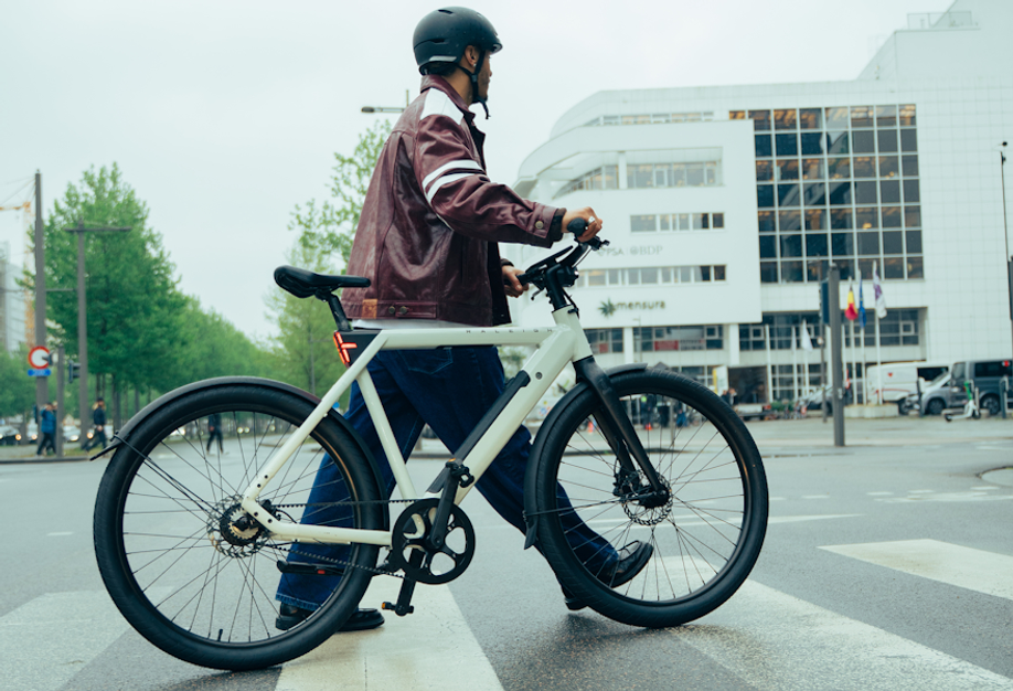 Man loopt met witte Rayleigh One urban e-bike over een zebrapad van een groot kruispunt voor een wit gebouw