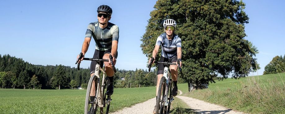 Man en vrouw gravelen op de Scott Addict Gravel fietsen over een gravelpad tussen de weilanden door met bomen erachter
