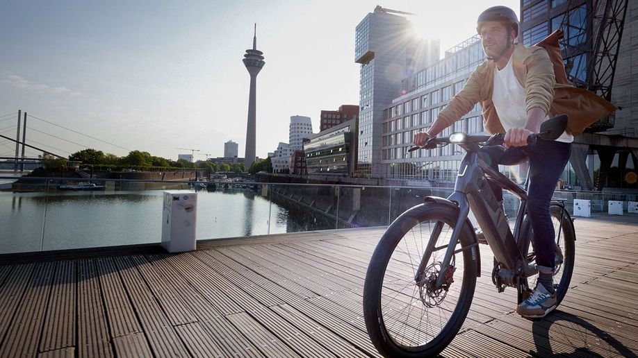 Man rijdt op zwarte Stromer speed pedelec over de houten planken van een grote brug in Rotterdam met gebouwen op achtergrond