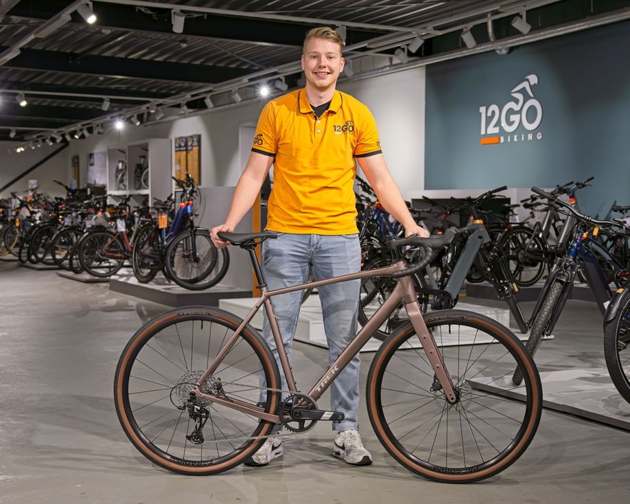 12GO Biking medewerker met Trek Checkpoint ALR 5 voor zich met 12GO Biking showroom achter zich
