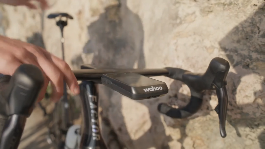 Wahoo Elemnt Ace op cockpitstuur voor een stenen muur waarvan de persoon de fiets gaat pakken