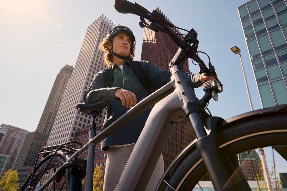 Man met helm leunt op fiets met zijn rechterarm met op de achtergrond veel hoge flatgebouwen en een stedelijke omgeving
