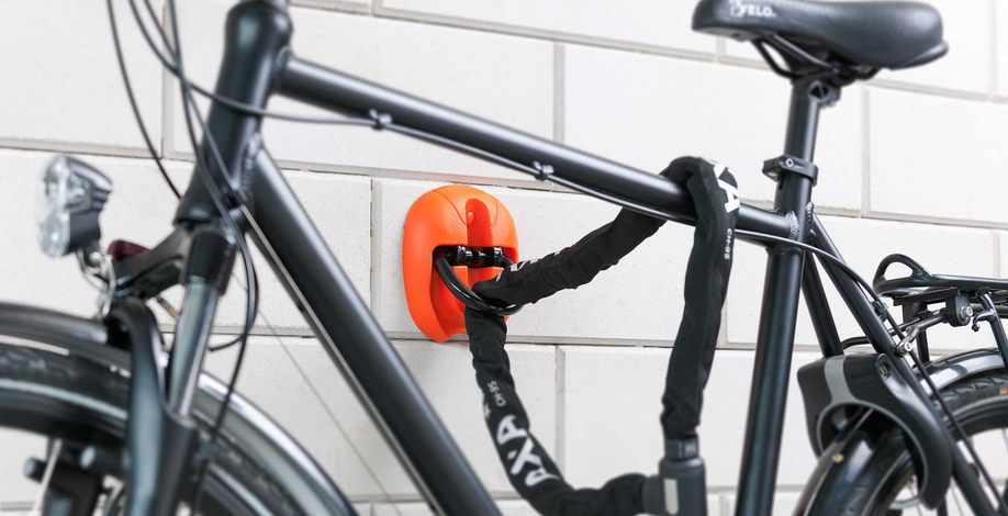 Zwarte fiets staat met AXA kettingslot van aan oranje muurbeugel die tegen aan de muur is geschroeft