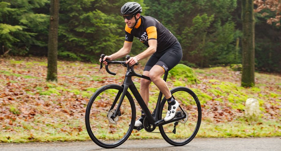 Fietser met 12GO Biking fietskleding en zwarte endurance racefiets die over het asfalt langs de bossen en bladeren rijdt