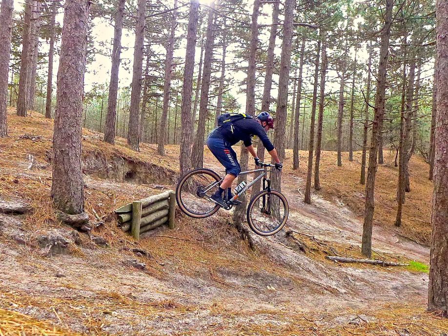 Man met halflange blauwe kleding springt met mountainbike van een hoogje af in een bos met veel droge bomen en ondergrond