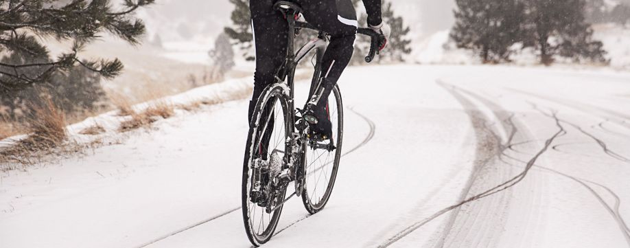 Man op zwarte racefiets met lange fietskleding die over een laag sneeuw op de weg heen fiets waarbij je meerdere sporen van andere fietsen en auto's ziet