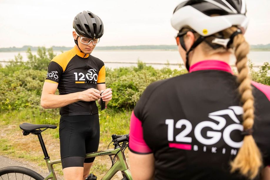 Man met groene Cannondale racefiets en 12GO Biking fietsshirt maakt een gel open terwijl er een vrouwelijke fietser met 12GO fietsshirt op de voorgrond staat