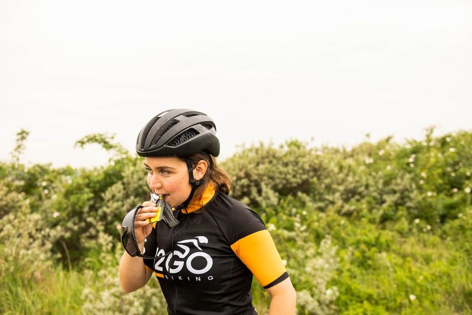 Vrouwelijke racefietser met 12GO Biking fietsshirt neem een hap van een PowerBar reep met groene struiken op de achtergrond