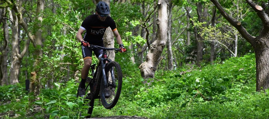 Mannelijke mountainbiker met 12GO Biking fietskleding die een sprong maakt over een bospaadje met veel groene bomen en struiken